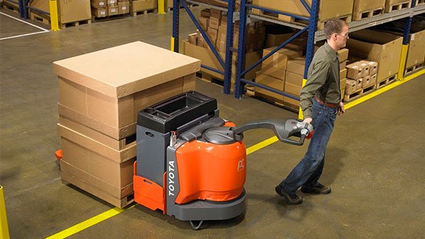 Man using a Toyota large electric walkie pallet jack to carry 2 large boxes across a warehouse 